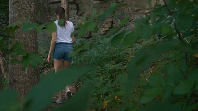 A young hipster woman leisurely hiking on a Tennessee park trail heading towards rocks and a cliff wearing shorts, wide shot, slow motion, cinematic