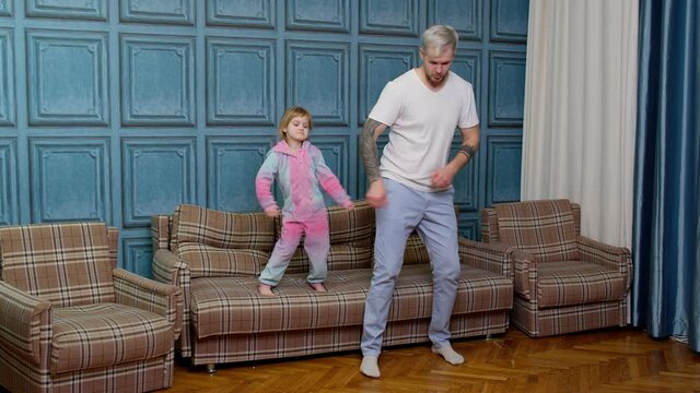 Young father babysiter and little kid daughter jumping dancing on sofa in modern house living room