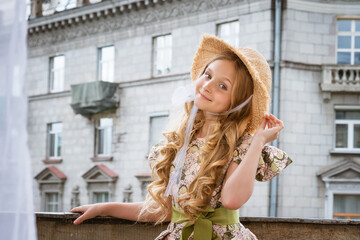 Little beautiful girl in dress and hat posing on the balcony against the background of the city