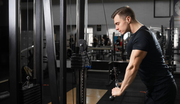 A male athlete in the gym stands behind a biceps and triceps machine and lowers the metal bars down.