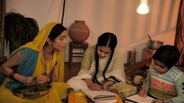 Rural housewife helping her children in studies while making dinner in the kitchen. Attractive village lady in a traditional saree cutting vegetables while her son and daughter doing homework - Vil...