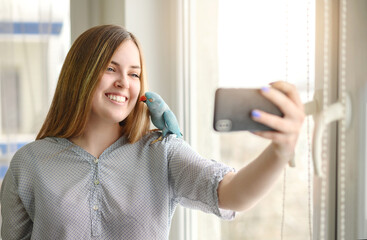 Pretty young woman with mobile phone by the window, with parrot on shoulder