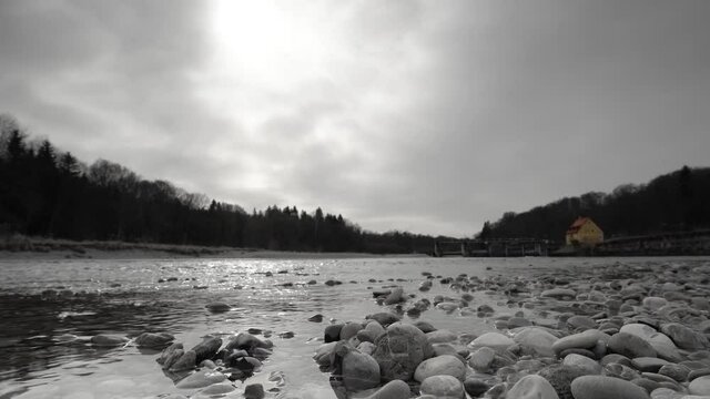 Black and white flowing river in front of a weir from lower, stony perspective with a colorkeyed yellow house in the background