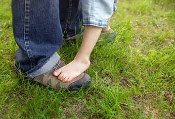 Fototapeta na wymiar Boy is happy with his first steps supported by his father on a summer meadow. The legs of the child boy stand on the feet of the father, dad supports the little son. Adorable feet on floor at nature.