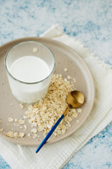 cup of milk on plate with scattered oatmeal and cereal flakes. beautiful golden spoon and white cotton napkin. vertical food content. plant-based alternative type of milk.