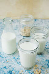 three cups milk and glass bottle with drink, scattered oatmeal on blue table. minimalism trendy food content. plant-based alternative type of milk. process of making oat milk at home, selective focus