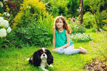 happy kid girl relaxing in summer garden and playing with her cavalier king charles spaniel dog