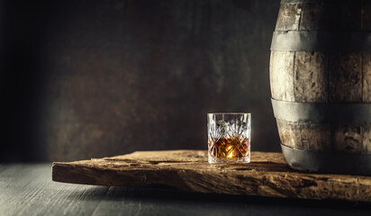 Glass of whisky or bourbon in ornamental glass next to a vinatge wooden barrel on a rustic wood and dark background - 420376561