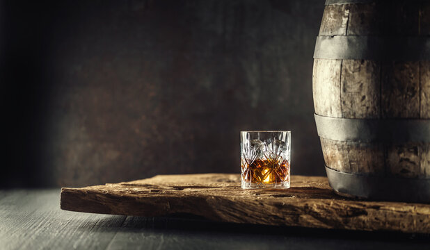 Glass of whisky or bourbon in ornamental glass next to a vinatge wooden barrel on a rustic wood and dark background