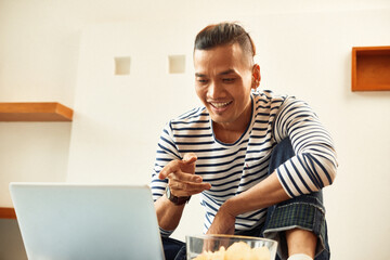 Smiling young Vietnamese man eating potato chips and watching comedy movie on laptop screen