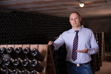 Male winemaker giving tour around winery, showing wine cellar with wine bottles racks