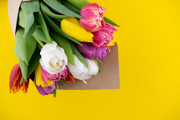 A bouquet of tulips wrapped in craft paper. Spring flowers. Gift delivery. Celebration. Copy space. Yellow, white, pink, purple buds. Yellow background. Postcard. Tulip flower.