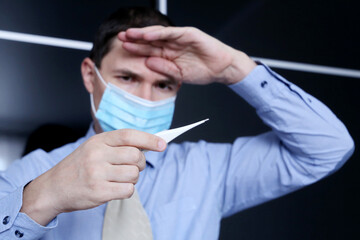 Worried man in face mask and office clothes looks at digital thermometer in hand. High body temperature, safety at work during coronavirus pandemic