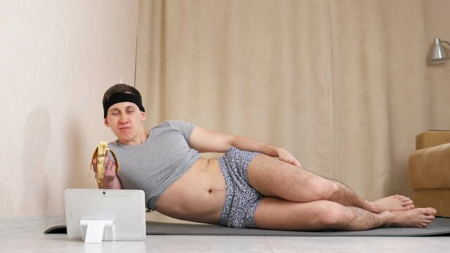 Feminine man eating banana and looking at tablet while lying on the floor.