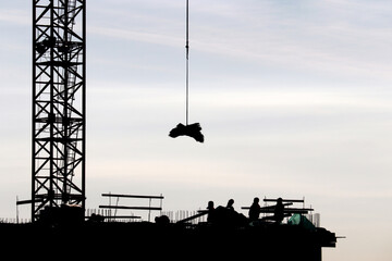 Silhouettes of workers and crane lifting the cargo on construction site against the sky. Housing...