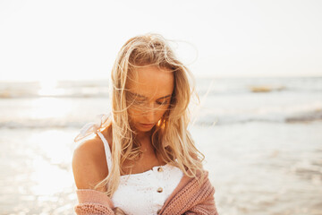 Headshot of beautiful blonde slim girlwearing white dress and cardigan on beach of sea or ocean against the sun. Youth, vacation, travel, fashion.