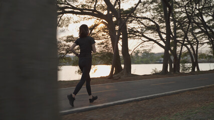 Obraz premium asian athlete woman in black sportswear and wireless headphone running in urban city trees park during sunset and bad cloudy weather. female jogging outdoor in evening. healthy workout.