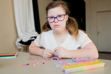 Cute teen girl stringing beads and making bracelet. Down syndrome kid crafting jewelry and threading bugle beads. Play and development of capacity disability children