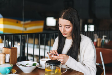 Attractive young brunette smiling woman in casual using mobile phone having brunch in the cafe