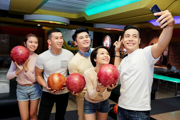 Group of Asian friends posing with balls at camera of mobile phone while playing bowling - obrazy, fototapety, plakaty