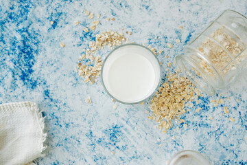 cup of milk and scattered oatmeal on blue table with white cotton napkin. analog type of milk. recipe for making oat milk at home.