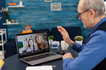 Fototapeta premium Senior man waving at daughter and niece during video conference. Happy grandfather saying hello in the course of online video confencere with family from living room.