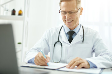 Cheerful doctor doing paperwork and using laptop in clinic