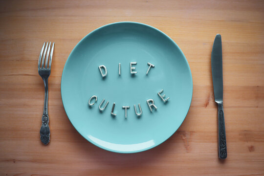 A blue plate, a fork and a knife on a wooden table. The letters on the plate say "diet culture" and stand for eating disorders