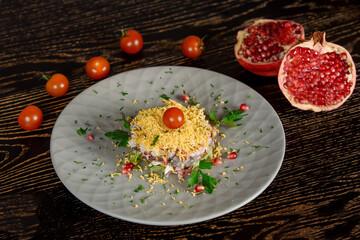 Puff salad with meat, vegetables, cheese, eggs, garnished with herbs and pomegranate and cherry tomatoes on a gray plate. Against the background of halves of pomegranate and cherry tomatoes
