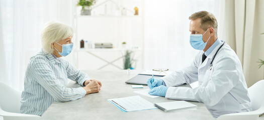 Fototapeta premium Doctor in medical mask sitting at the table and talking with old lady