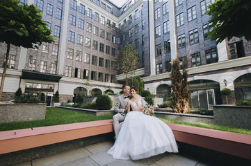 Stylish groom in a gray suit and a bride in a white dress are hugging in a park with green plants, on the background of buildings in the city, sitting on a wooden bench. Wedding portrait.