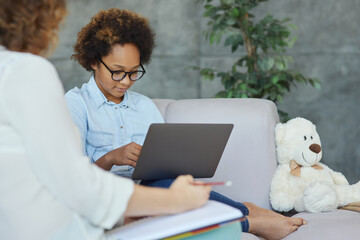 Cute teen girl in glasses listening to her female teacher, using laptop for studying while having a lesson, sitting on the couch at home