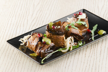 Eggplant rolls, garnished with herbs, pomegranate, suluguni cheese. Georgian appetizer on a black square plate on a white wooden table.