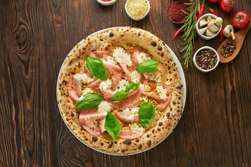 Delicious homemade neapolitan pizza with ham, mozzarella cheese, fresh basil and food ingredients served on wooden table, fast food italian kitchen and eating concept
