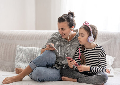 A young mother and her daughter are listening to music on their phones while sitting on the couch at home.