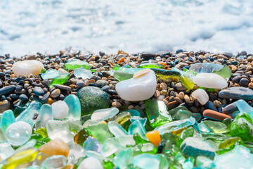 Natural polish textured sea glass and stones on the seashore. Azure clear sea water with waves. Green, blue shiny glass with multi-colored sea pebbles close-up. Beach summer background