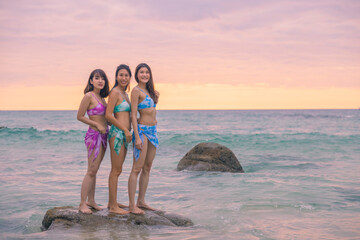 Three young women friends happily wear this green, blue, purple bikini standing on the rocks at Nai Thon Beach, Phuket, Thailand. Portrait of happy young woman smiling at sea. Concept of Travel