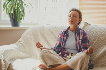 Fototapeta premium Young woman sitting on couch at home meditating with closed eyes, wearing casual cloth. Doing yoga exercises, breathing practices to calm down mind, relieve tension and stress. Lotus pose. Copy space