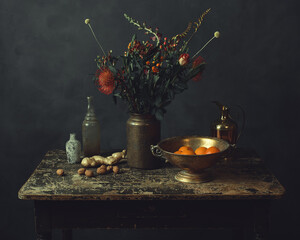 Old worn wooden table topped with a vase with flowers, ginger and walnuts, a bronze bowl with oranges, a pitcher and two glass bottles.