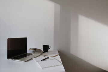 Laptop on table with coffee cup, paper sheet against white wall. Minimalist home office workspace.