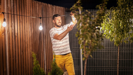 Party time in backyard with happy man hanging string lights in trees - 420417939