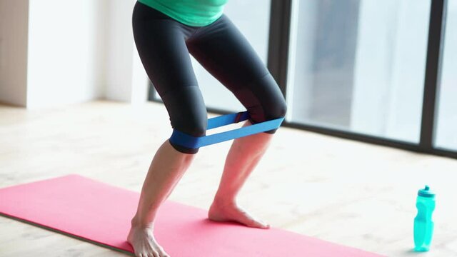 Keep fit. Close up of sportive woman exercising with rubber band, training her legs while working out on fitness mat at home in the living room