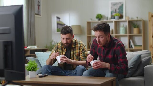 Two young cute men Caucasian eating a Chinese noodle sitting in a living room on a couch and watch TV