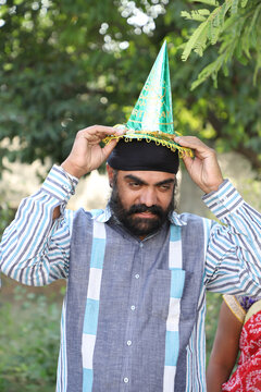 Middle-aged Indian Man With A Turban On His Head Trying To Wear A Paper Hat For A Birthday