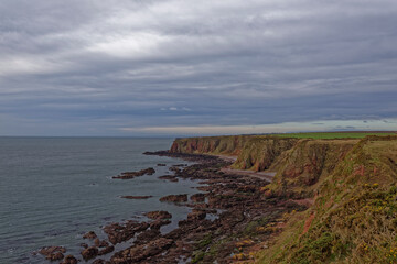 Shingle Beaches, rocky outcrops and Red Sandstone Cliffs with Farmland above seen from the Angus Coastal Footpath under a dark sky.