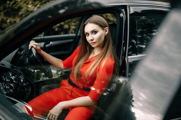 Fototapeta na wymiar A beautiful young girl in a red overalls sits behind the wheel of a black car on an empty road in the forest