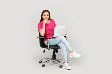 Young woman with laptop sitting in comfortable office chair on grey background