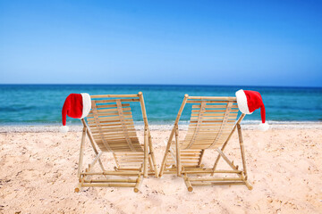 Wooden deck chairs and Santa Claus hats on beach near sea. Christmas vacation