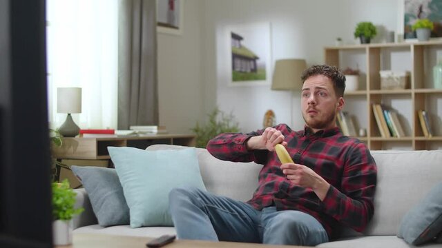 Young handsome man eats banana and watching tv at home in the living room.