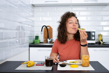 Obraz na płótnie Canvas Young brunette woman enjoying breakfast in white kitchen at home,sitting at table chewing healthy bread with avocado and peanut butter looking at camera with joy.Morning routine.Vegetarianism concept